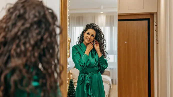 Frau mit dunklen Haaren steht vor dem Spiegel, macht sich ihre Ohrringe rein, trägt ein grünes Wickelkleid - Foto: iStock/ supersizer