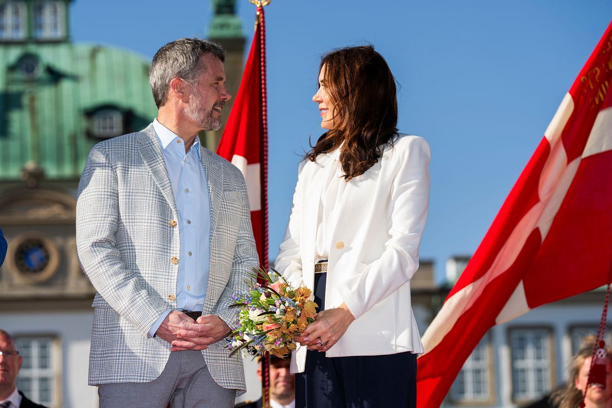 Frederik und Mary blicken sich vor Schloss Fredensborg ins Gesicht, im Hintergrund wehende Dänemarkflaggen und ein blauer Himmel.