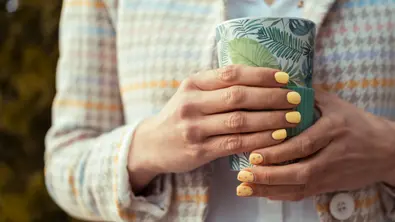Frau mit patellgelben „Eierpunsch-Nägeln“ hält Becher in der Hand. - Foto: iStock