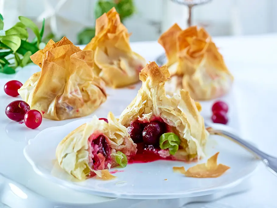4 goldbraune Cranberrypäcken, davon ein aufgeschnittenes im Vordergrund auf einem weißen Teller angerichtet.