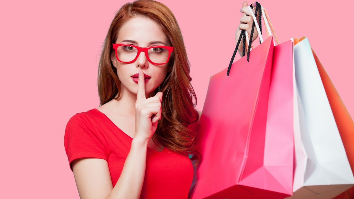 Frau mit roter Brille und roten haare hält Shoppingtüten hoch und hält sich der rechten Zeigefinger vor den Mund