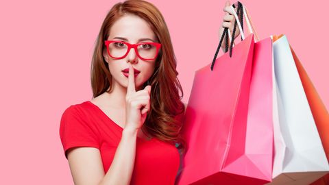 Frau mit roter Brille und roten haare hält Shoppingtüten hoch und hält sich der rechten Zeigefinger vor den Mund - Foto: iStzock/Massonstock