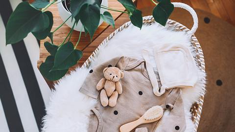 Ein Teddybär, ein Strampler und Babysöckchen drapiert in einer Kinderwiege, alles in neutralen Farbtönen gehalten - Foto: iStock/NataliaDeriabina