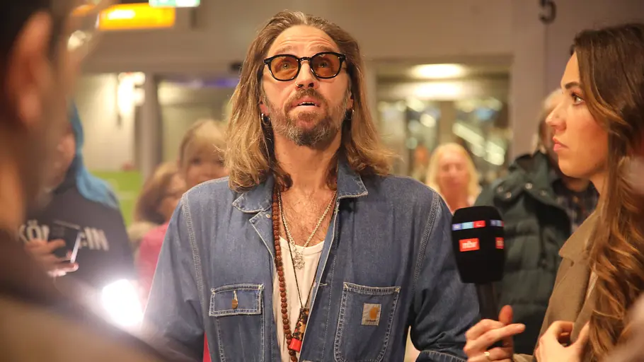 Gil Ofarim. Gil Ofarim bei der Ankunft am Flughafen, er trägt eine Sonnenbrille und blickt nach oben, rechts neben ihm steht eine Reporterin mit einem Mikrofon
