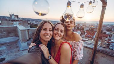 Drei Frauen posieren unter einer Glühbirnen-Lichterkette. - Foto: iStock/urbazon