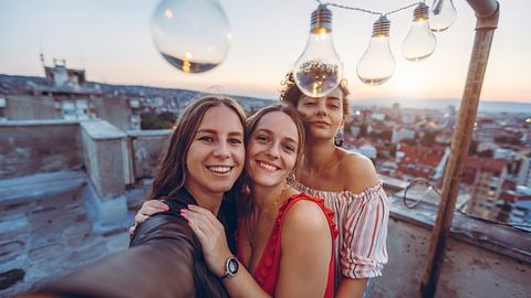 Drei Frauen posieren unter einer Glühbirnen-Lichterkette. - Foto: iStock/urbazon