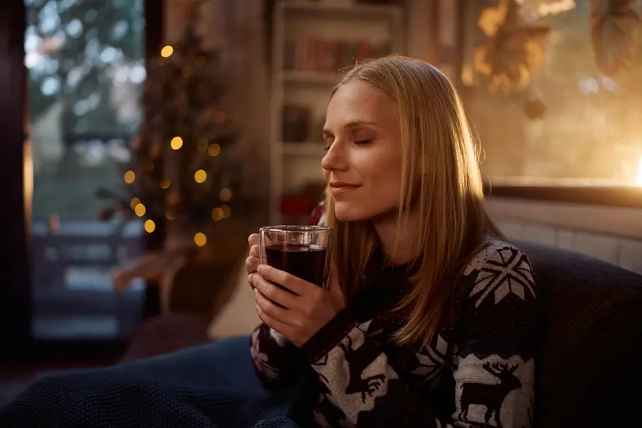 Wie lange ist Glühwein ungeöffnet haltbar Eine Frau sitzt mit einem Glas Glühwein in der Hand in ihrem Wohnzimmer, im Hintergrund leuchtet ein geschmückter Weihnachtsbaum