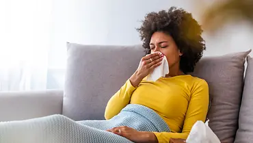 Frau liegt erkältet auf dem Sofa unter einer Decke und schnäuzt ins Taschentuch (Themenbild) - Foto: dragana991/iStock Frau liegt erkältet auf dem Sofa unter einer Decke und schnäuzt ins Taschentuch (Themenbild) - Foto: dragana991/iStock