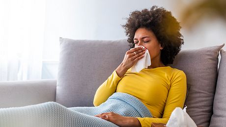 Frau liegt erkältet auf dem Sofa unter einer Decke und schnäuzt ins Taschentuch (Themenbild) - Foto: dragana991/iStock