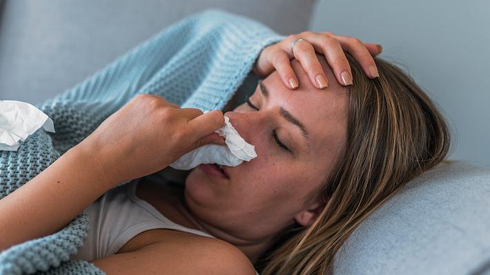 Frau liegt verschnupft auf dem Sofa und schnäuzt die Nase, neben ihr liegen zerknüllte Taschentücher (Themenbild) - Foto: ljubaphoto/iStock