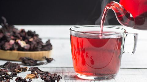 Hibiskustee wird in ein durchsichtiges Henkelglas gegossen und dampft, daneben liegen getrocknete Hibiskusblüten auf einem Tisch (Themenbild) - Foto: mescioglu/iStock