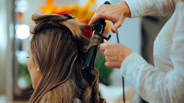 Frau mit braunem Haar bekommt Locken mithilfe eines Lockenstabs.  - Foto: nicoletaionescu/iStock (Themenbild)