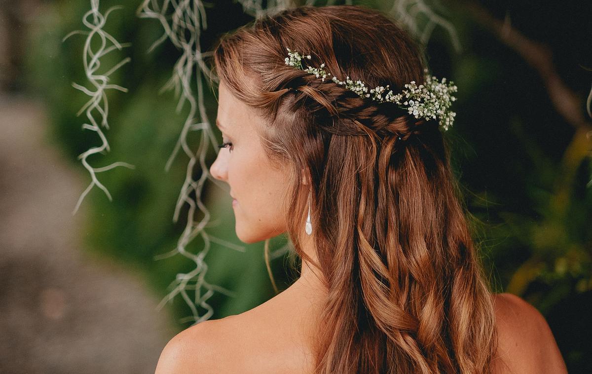 Braut trägt ihre langen Haare halboffen. Ein Blumengeflecht am Hinterkopf verschönert die Frisur.