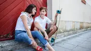 Mutter sitzt mit ihrem Teenager-Sohn mit Skateboard im Arm auf einer Bordsteinkante und unterhält sich mit ihm - Foto: PR/Canva.de