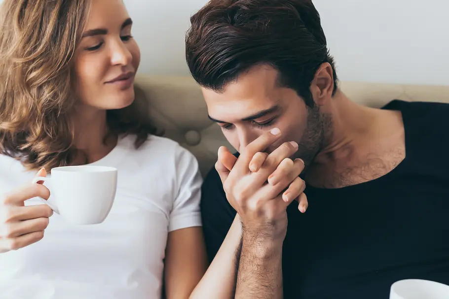 Erotische Liebesgeschichte Porträt eines verliebten Paares: Ein Mann küsst die Hand einer Frau während sie auf einem Sofa sitzen. Sie hält in der anderen Hand eine Tasse Kaffee und lächelt.