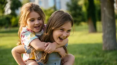 Älteres Mädchen trägt jüngeres Mädchen auf dem Rücken und beide lachen - Foto: Drazen_/ iStock