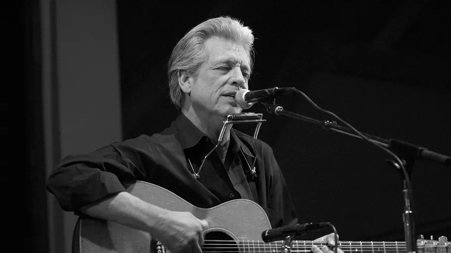 John Hammond singt sitzend in ein Mikrofon und spielt Gitarre. John Hammond singt sitzend in ein Mikrofon und spielt Gitarre.