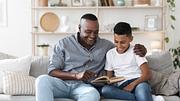 Ein Vater sitzt gemeinsam mit seinem Sohn auf einem Sofa und sie lesen gemeinsam ein Buch - Foto: iStock/Prostock-Studio