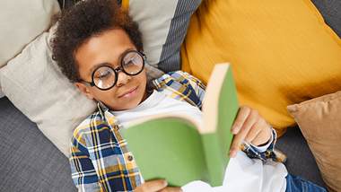 Ein Junge mit Brille liegt auf dem Sofa und liest ein Buch - Foto: iStock/SeventyFour