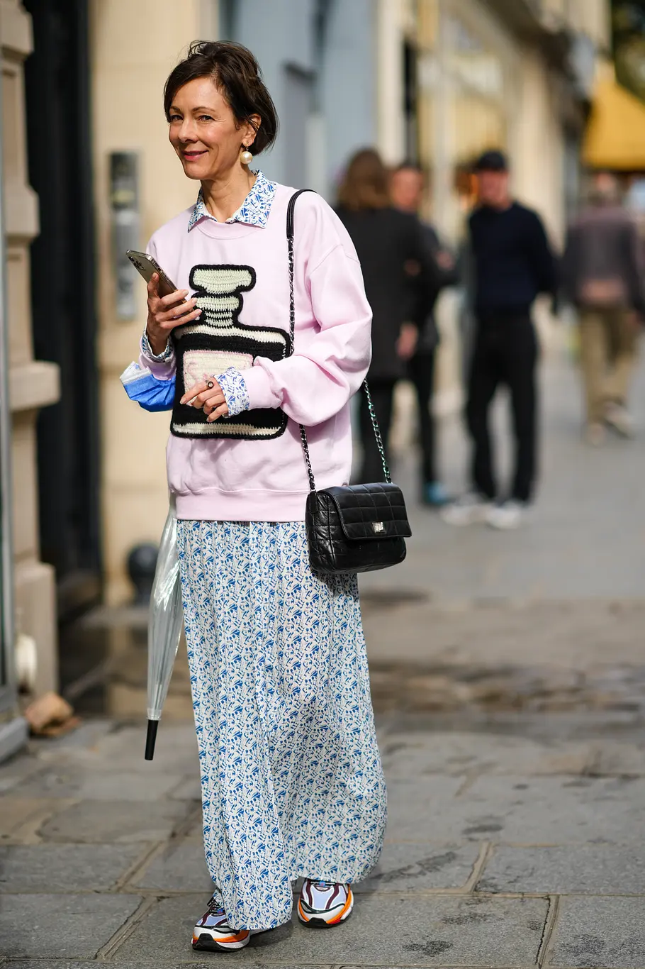 A guest wears large pearls pendant earrings, a white with blue print pattern shirt flowing jumpsuit, a pale pink with a black and white embroidered large perfume bottle pattern sweater