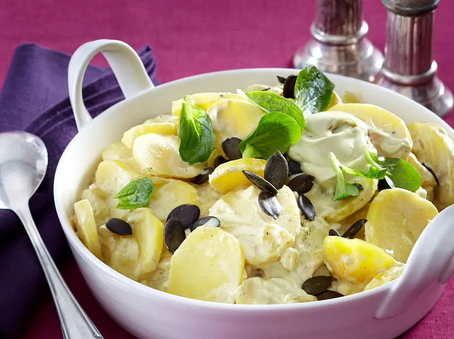 Auf einem gedeckten Tisch steht eine weiße Porzellanschüssel mit cremigem Kartoffelsalat, der mit Feldsalat und Kürbiskernen garniert ist