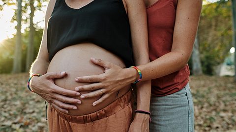 Nahaufnahme eines lesbischen Paares, das ihren Schwangerschaftsbauch in einem Park hält - Foto: Wirestock/iStock