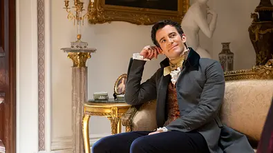 Ein Mann in historischer Kleidung sitzt entspannt auf einem eleganten Sofa in einem prunkvollen Salon mit goldenen Möbeln und klassischer Dekoration. - Foto:  LIAM DANIEL/NETFLIX 