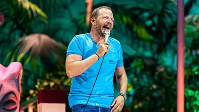 Ein Mann steht auf einer Bühne vor tropischer Kulisse und hält ein Mikrofon in der Hand. Er trägt ein blaues T-Shirt mit der Aufschrift „Mario Barth Tour 2018“ sowie Jeans und lacht mit weit geöffnetem Mund, als würde er gerade einen Witz erzählen. Im Hintergrund sind grüne Pflanzen, bunte Bühnenbeleuchtung und ein dekoratives Element zu sehen. - Foto: RTL / Sebastian Drüen