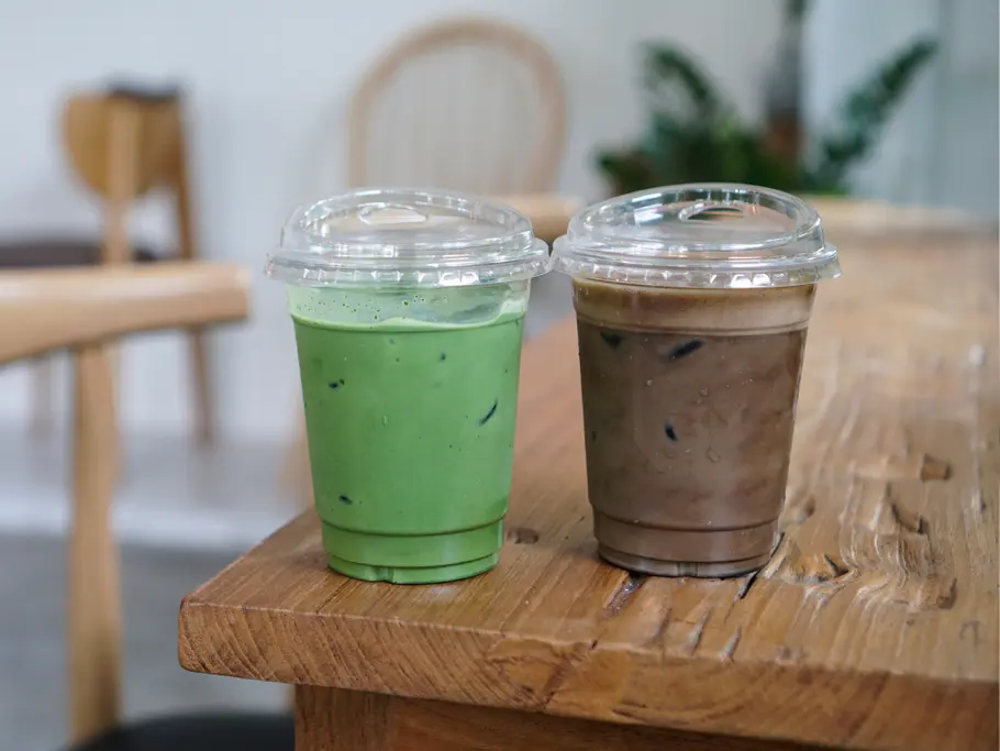 Matcha vs. Hojicha Ein Plastikbecher mit grünem Matcha neben braunem Hojicha vor Café-Kulisse