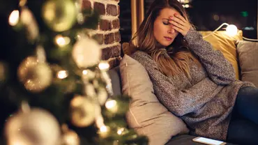Junge Frau sitzt gestresst auf dem Sofa vor einem weihnachtlich geschmückten Hintegrund - Foto: i-Stock/svetikd
