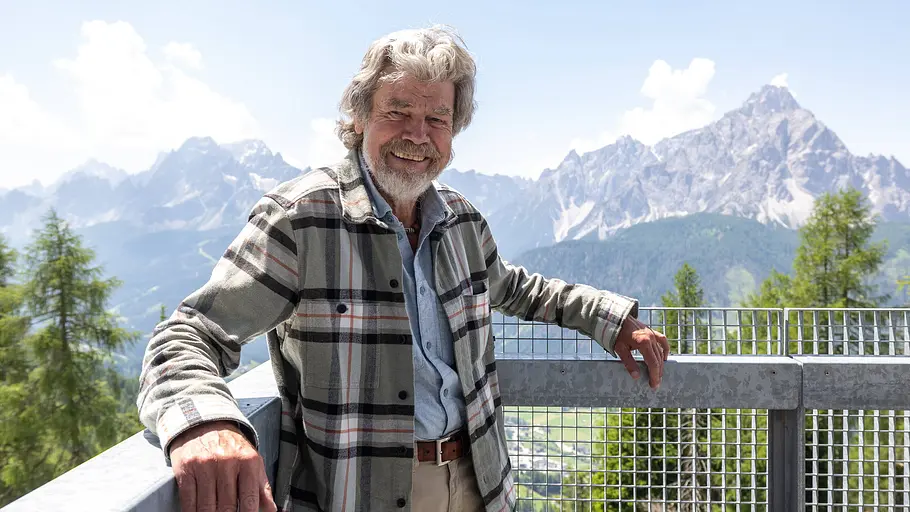 Reinhold Messner vor beeindruckender Alpenkulisse Reinhold Messner steht lächelnd auf einer Aussichtsplattform mit Bergpanorama im Hintergrund.