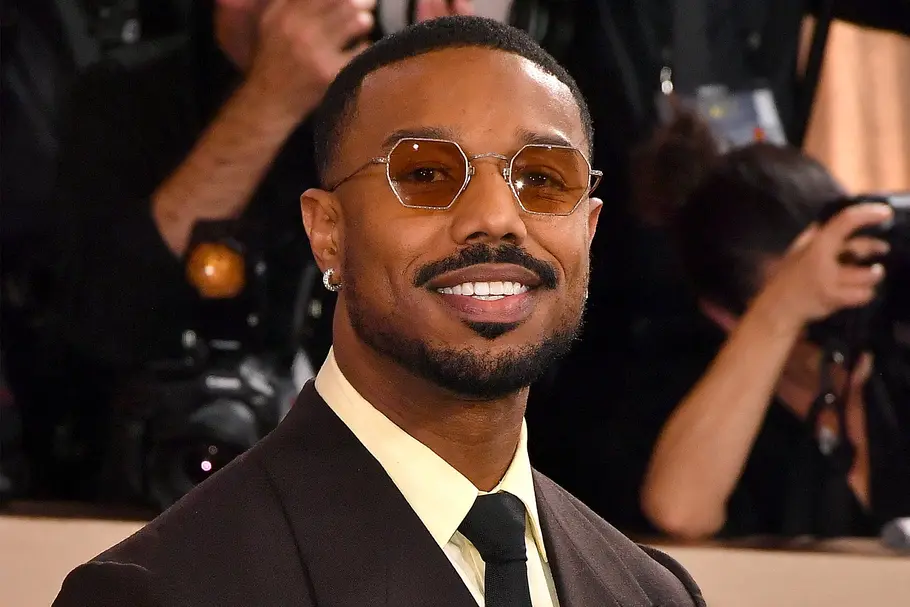 Der Schauspieler Michael B. Jordan steht auf dem roten Teppich, trägt einen dunkelbraunen Anzug, eine Sonnenbrille und lächelt in die Kamera.