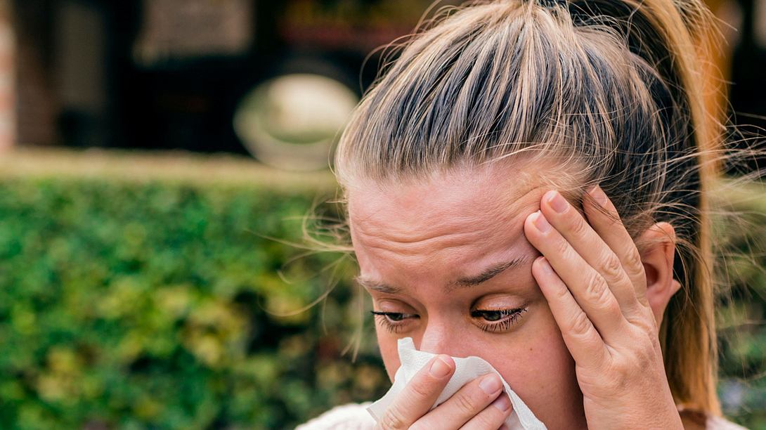 Frau mit allergischem Augenbrennen putzt sich die Nase und hält sich schmerzenden Kopf im Garten. - Foto: dragana991/iStock