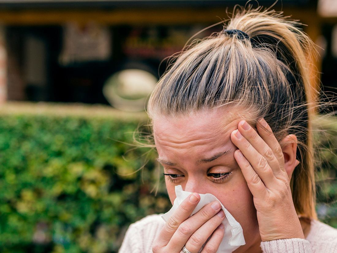 Frau mit allergischem Augenbrennen putzt sich die Nase und hält sich schmerzenden Kopf im Garten.
