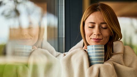 Frau setzt in Decke gehüllt und hat eine Tasse in der Hand - Foto: iStock/nortonrsx