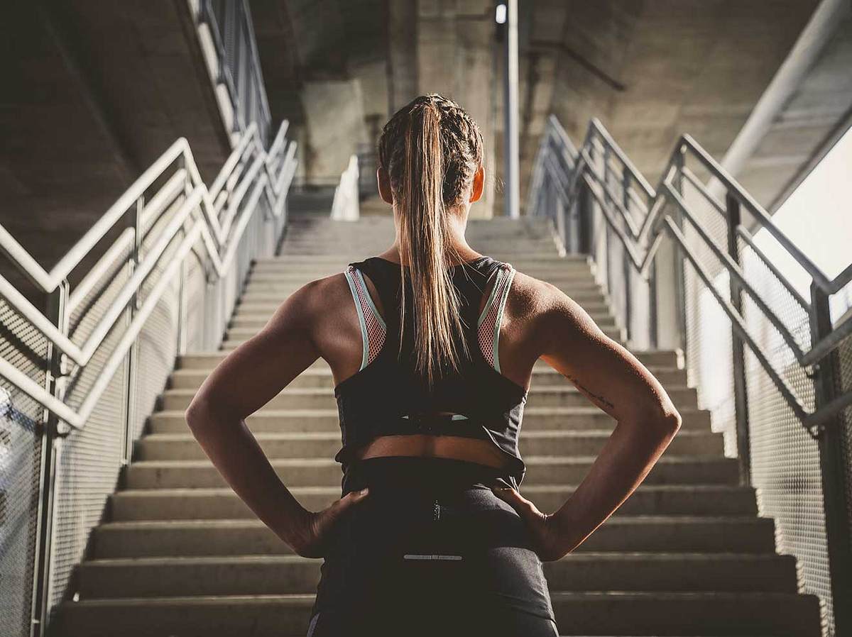 Frau in Sportoutfit steht vor Treppe und hat die Hände in die Hüften gestemmt (Themenbild)