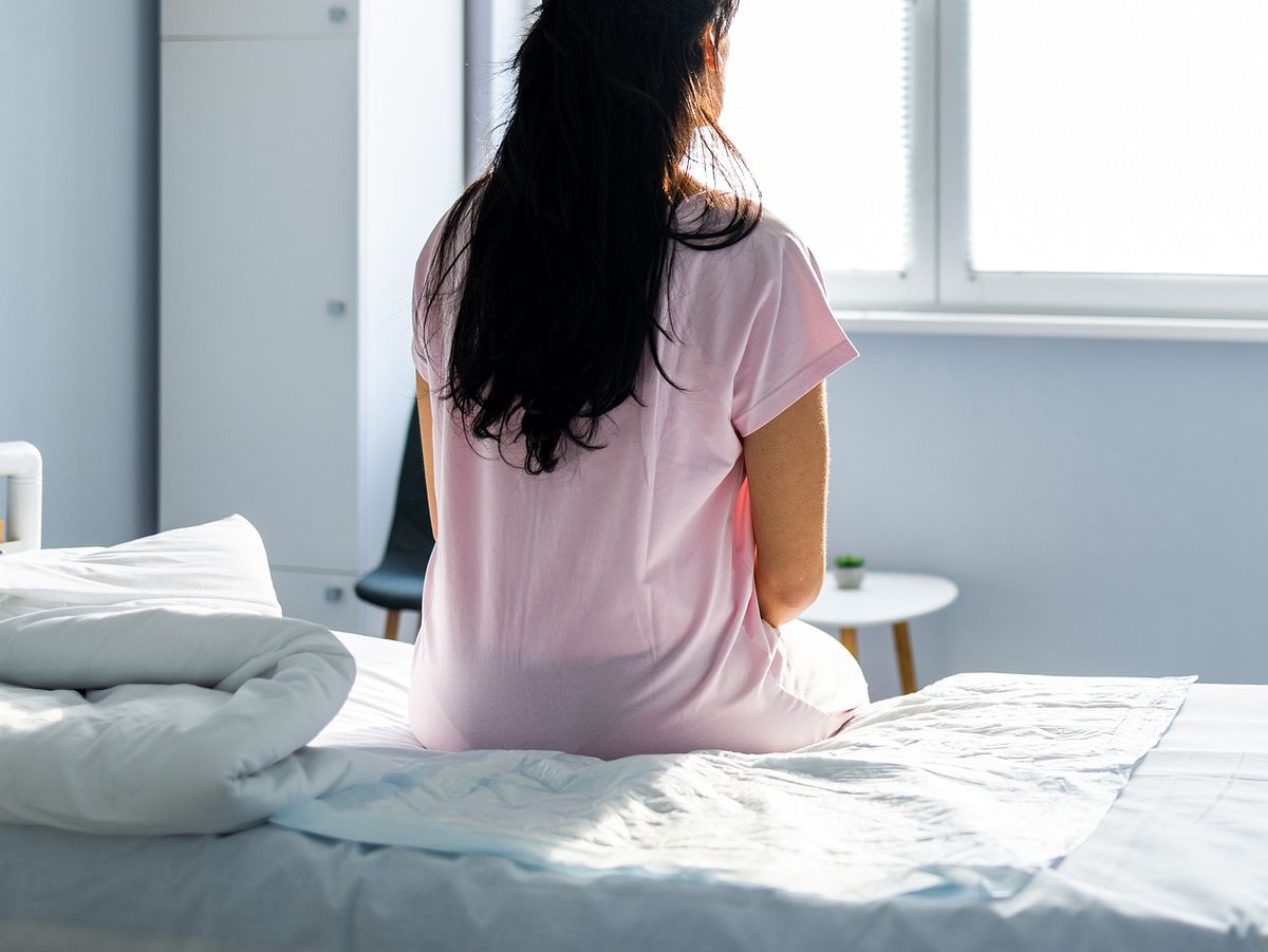 Frau sitzt traurig auf einem Krankenhausbett - der Rücken ist dem Betrachter zugewandt.
