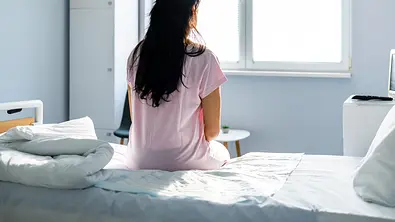 Frau sitzt traurig auf einem Krankenhausbett - der Rücken ist dem Betrachter zugewandt. - Foto: ljubaphoto / iStock