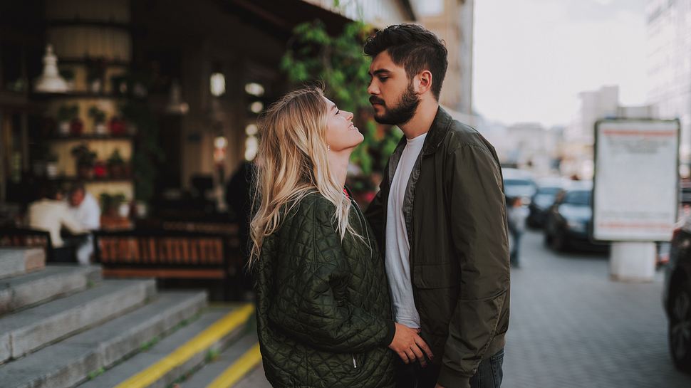 Mann und Frau stehen in der Stadt. Die Frau lächelt und richtet sich zum Mann. Der Mann guckt ernst und wirkt distanziert. (Themenbild) - Foto: YakobchukOlena/iStock