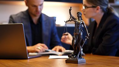 Justizia-Statue mit Waage und Schwert, dahinter Kolleg*innen beim Diskutieren (Themenbild) - Foto: Vladislav Stepanov/iStock