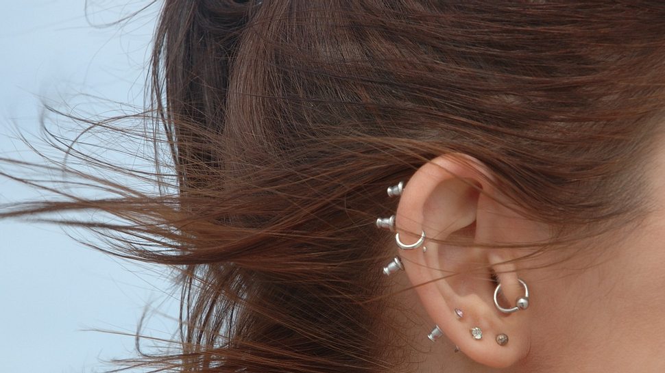 Ohr einer Frau mit vielen Piercings und Steckern im Ohr, die langen Haare wehen im Wind (Themenbild) - Foto: EyeJoy/iStock