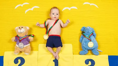 Baby liegt mit Medaille um den Hals auf einer Decke, die aussieht wie ein Siegerpodest. Es ist rechts und links flankiert von Kuscheltieren, die ebenfalls Medaillen tragen. - Foto: Elena Bessonova / iStock