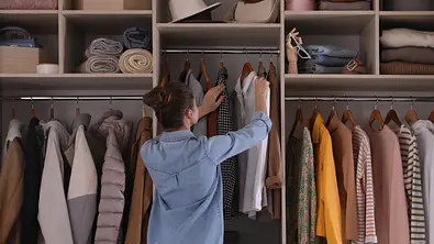 Frau mit Bluse und Dutt vor einem offenen Kleiderschrank bei der Outfitwahl - Foto: iStock / Liudmila Chernetska