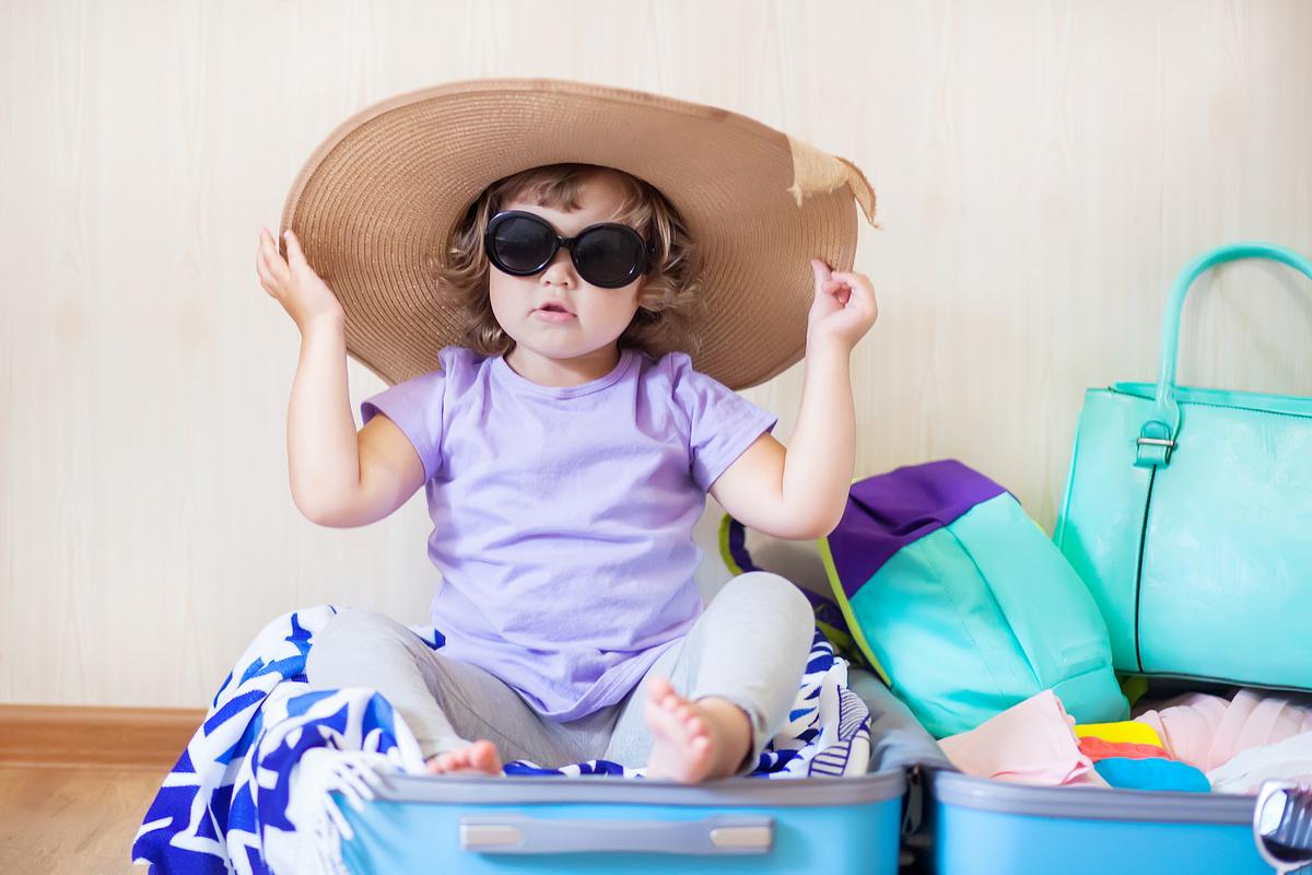 Kleinkind sitzt im geöffneten Koffer mit Sonnenbrille und Hut