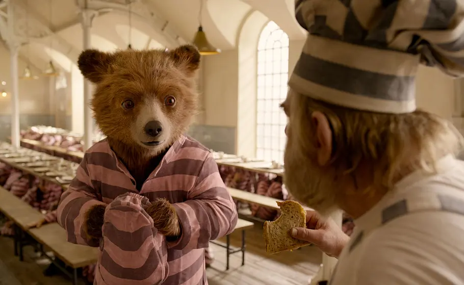 Paddington 2 Paddington hat ein rosa gestreiftes Outfit an. Ein Bäcker isst ein Brot und meckert ihn an.