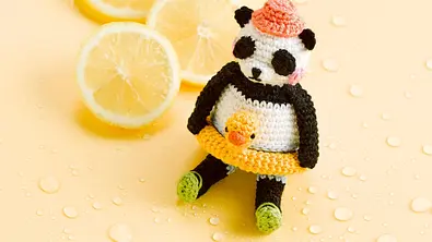 Ein kleiner und süßer gehäkelter Panda mit gelbem Quietschenten-Schwimmring, rosa Sonnenhut und grünen Badeschuhen.  - Foto: Frechverlag