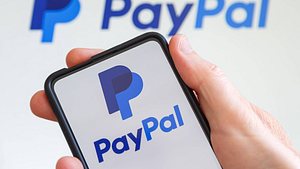 Eine Hand hält ein Handy, auf dem das PayPal-Logo abgebildet ist. Im Hintergrund sieht man ebenfalls das PayPal-Logo. - Foto: IMAGO / Zoonar