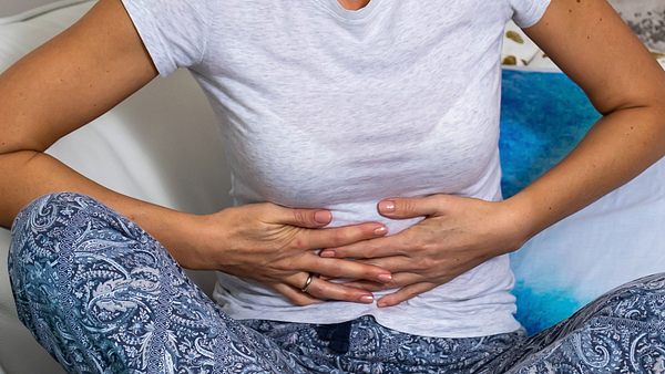 Frau sitzt mit Unterleibsschmerzen auf dem Sofa. - Foto: RealPeopleGroup / iStock