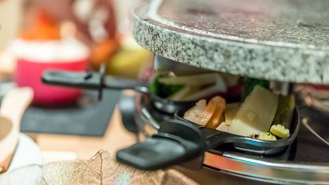 Raclette Grill mit Pfännchen und Zutaten auf dem Esstisch. - Foto: yevtony/iStock