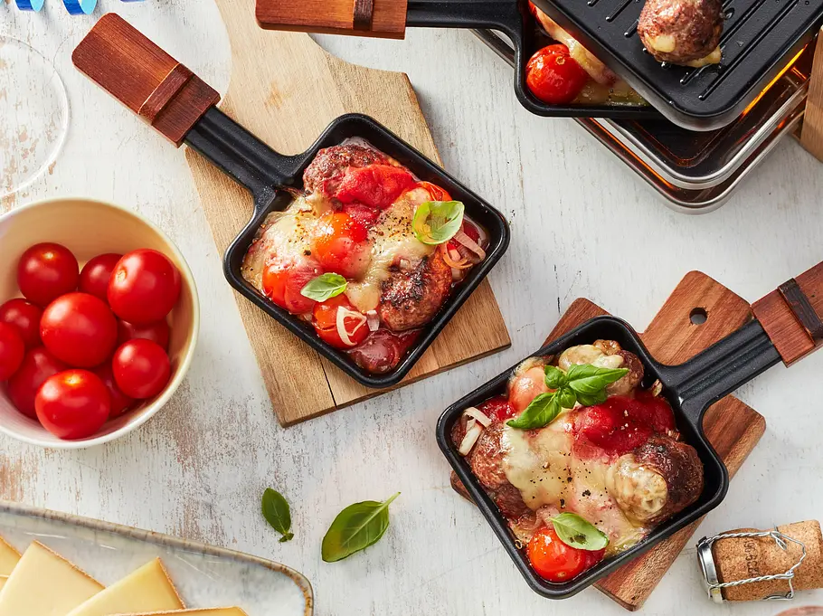 Raclette-Idee Weihnachten: Auf einem Tisch liegen zwei kleine Holzbretter, auf jedem steht ein Raclettepfännchen mit Hackbällchen, Tomaten, Raclettekäse und frischem Basilikum